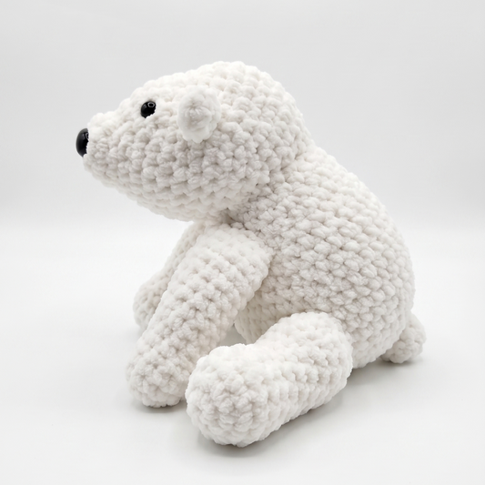 A white crocheted polar bear toy with black embroidered eyes and nose, side sitting on a light gray studio background.