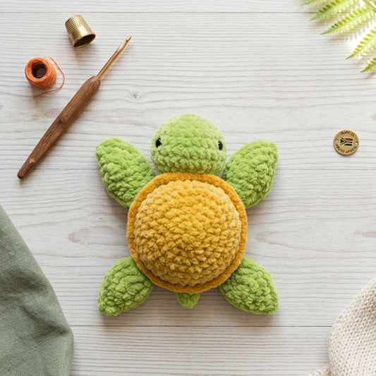 A crocheted turtle toy with a green body and yellow shell, styled on a light wood surface with crochet tools, fabric, and a fern leaf.