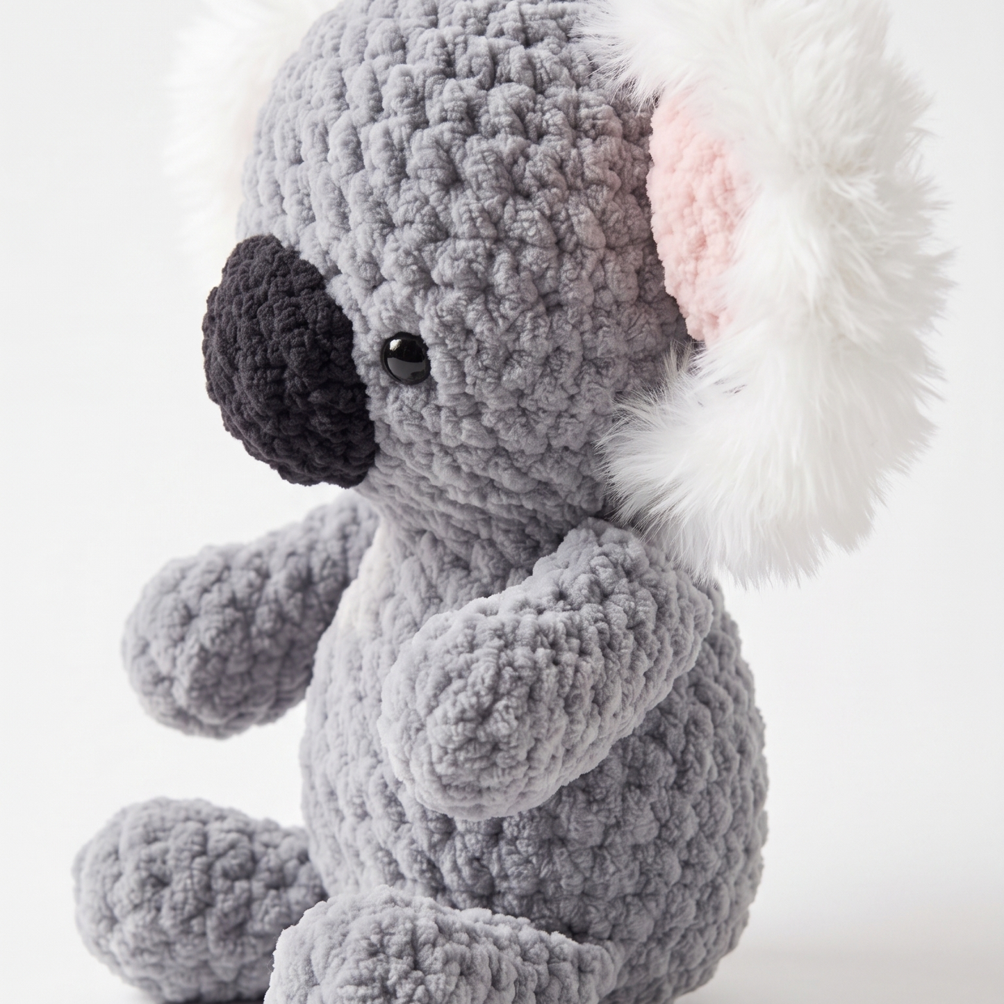 A crocheted koala plush toy in gray with white fluffy ears, a black nose, and a pink inner ear, photographed on a clean light gray background.