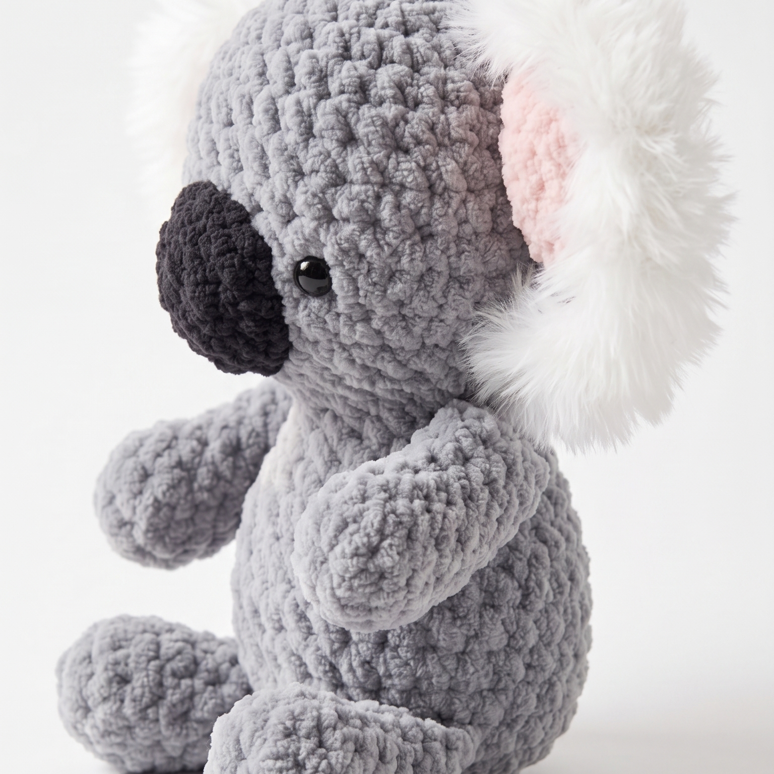 A crocheted koala plush toy in gray with white fluffy ears, a black nose, and a pink inner ear, photographed on a clean light gray background.