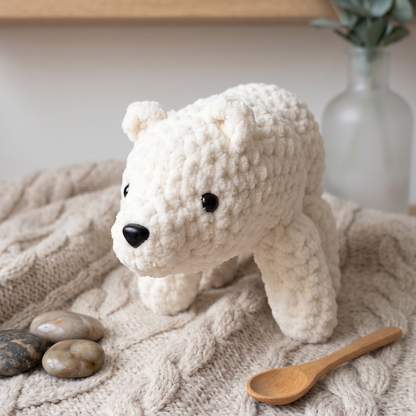 A small crocheted polar bear toy in off-white sits on a beige cable-knit blanket, with a wooden spoon, smooth stones, and a glass vase with greenery in the background.