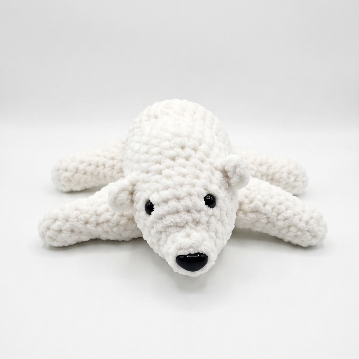 A white crocheted polar bear toy with black embroidered eyes and nose, lying on a light gray studio background.