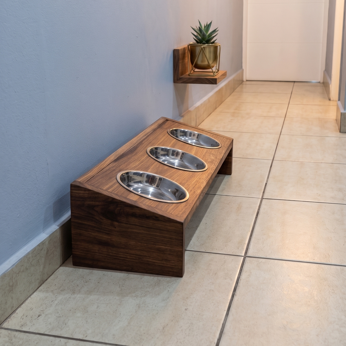 A wooden elevated pet feeder stand with three stainless steel bowls, placed on a tiled floor in a hallway next to a small shelf with a plant.