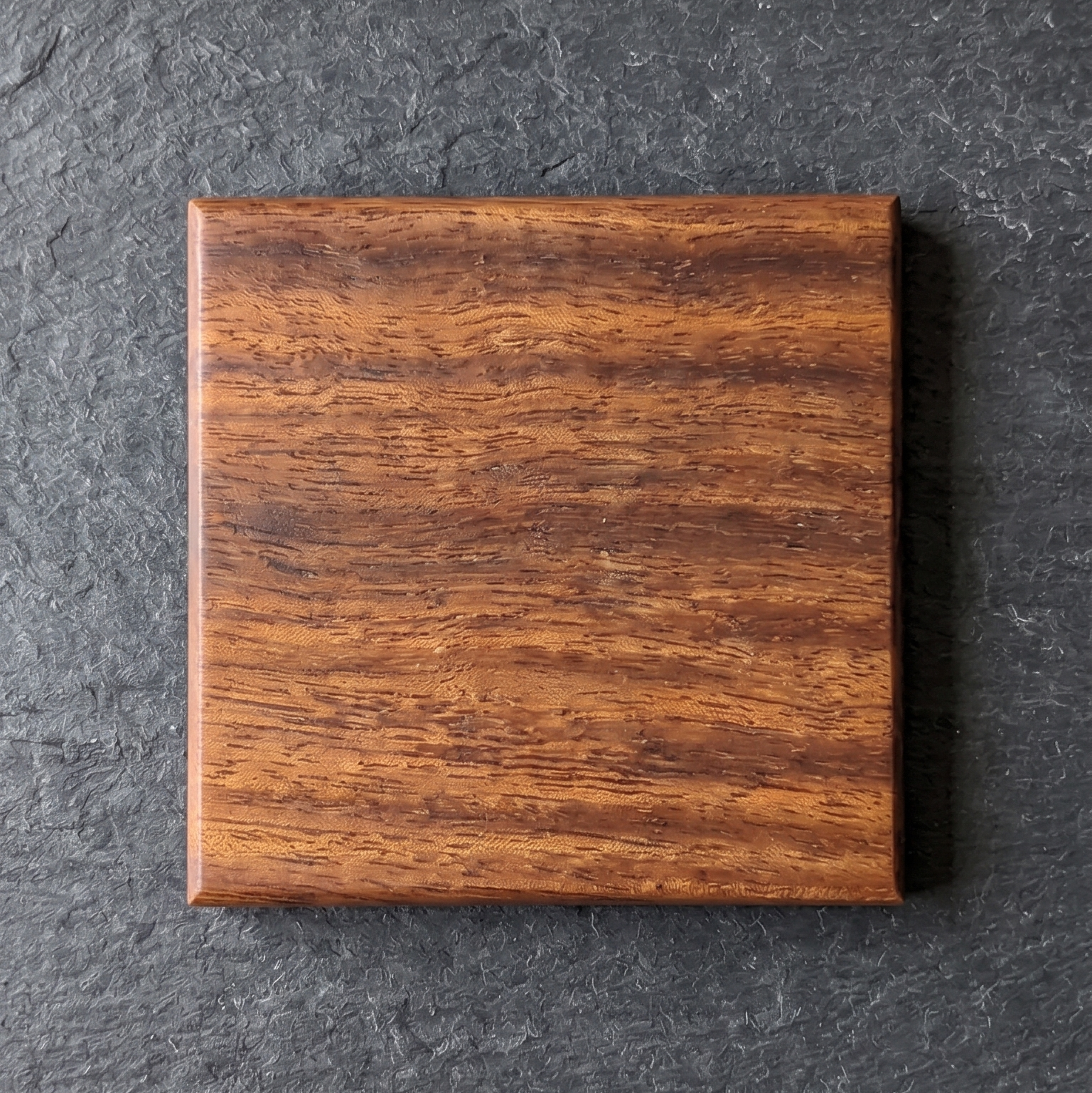 A square African Walnut coaster with a hardwax oil finish, centered on a dark gray slate background